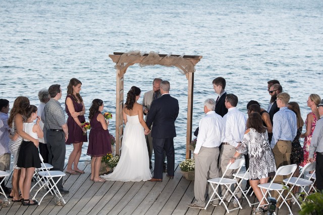 A couple exchanging vows on a dock by the ocean, surrounded by guests and decorated with flowers and drapery.