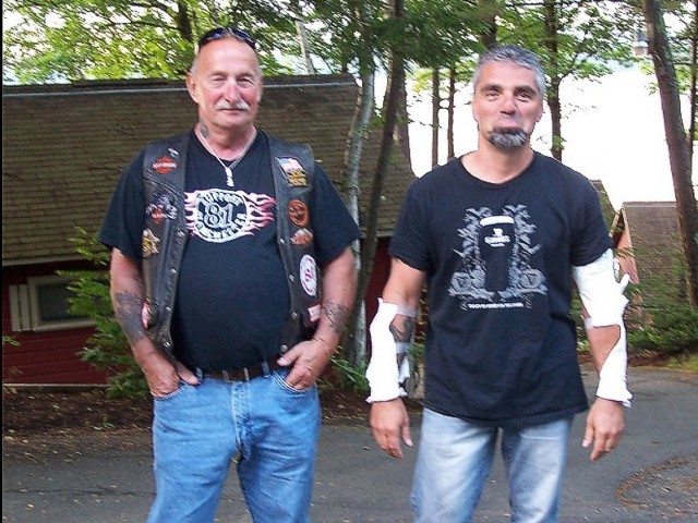 Two men standing side by side in front of a house, smiling and dressed casually.