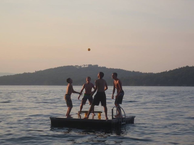 Four men are playing with a ball on a raft floating in the water, enjoying a sunny day outdoors.