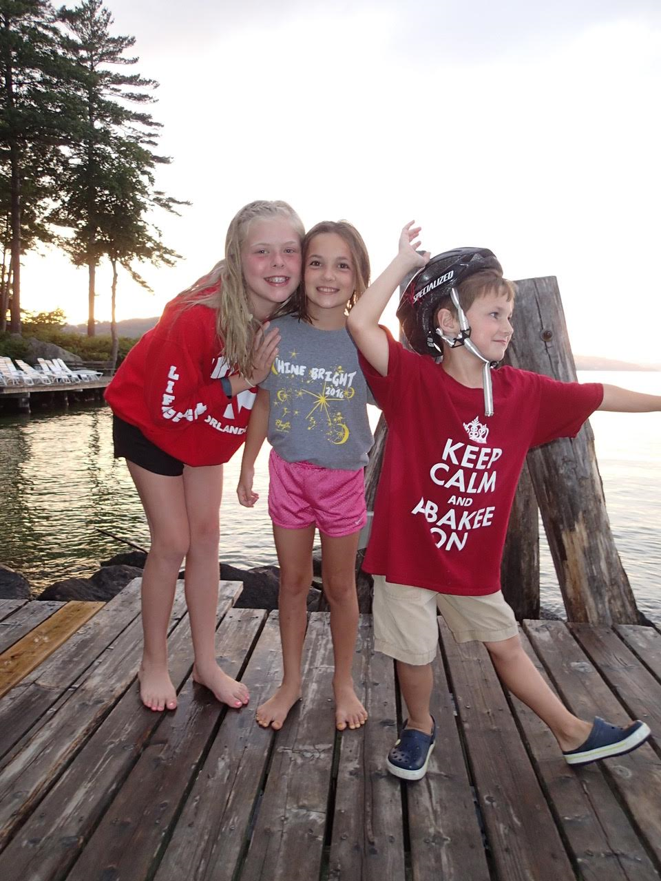 Three children in red shirts standing together on a wooden dock, smiling and looking out over the water.