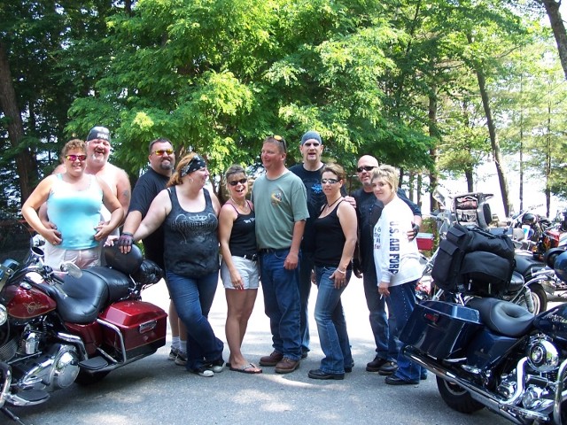 A group of people standing beside various motorcycles in an outdoor setting, smiling and engaging with each other.
